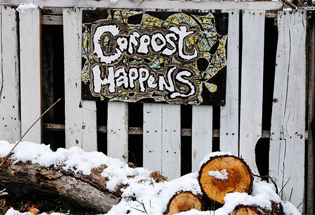 A sign on a fencepost that says 'Compost happens' while there are logs cover with snow underneath the sign.